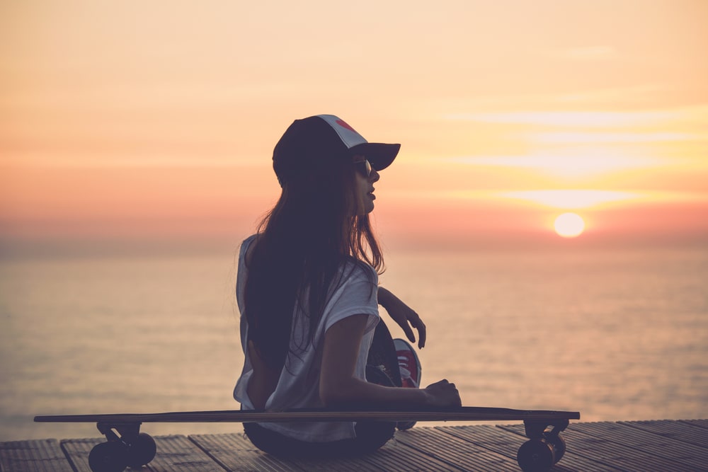 A skater girl watching the sunset and sitting by the dock, leaning on her skateboard.