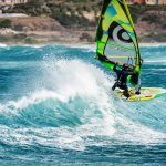 A windsurfer wearing the necessary windsurfing protective gear, riding the rough waves with his yellow sailboard.