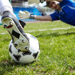 A soccer player wearing soccer cleats, about to kick the ball toward the goal, with the goalie blocking it.